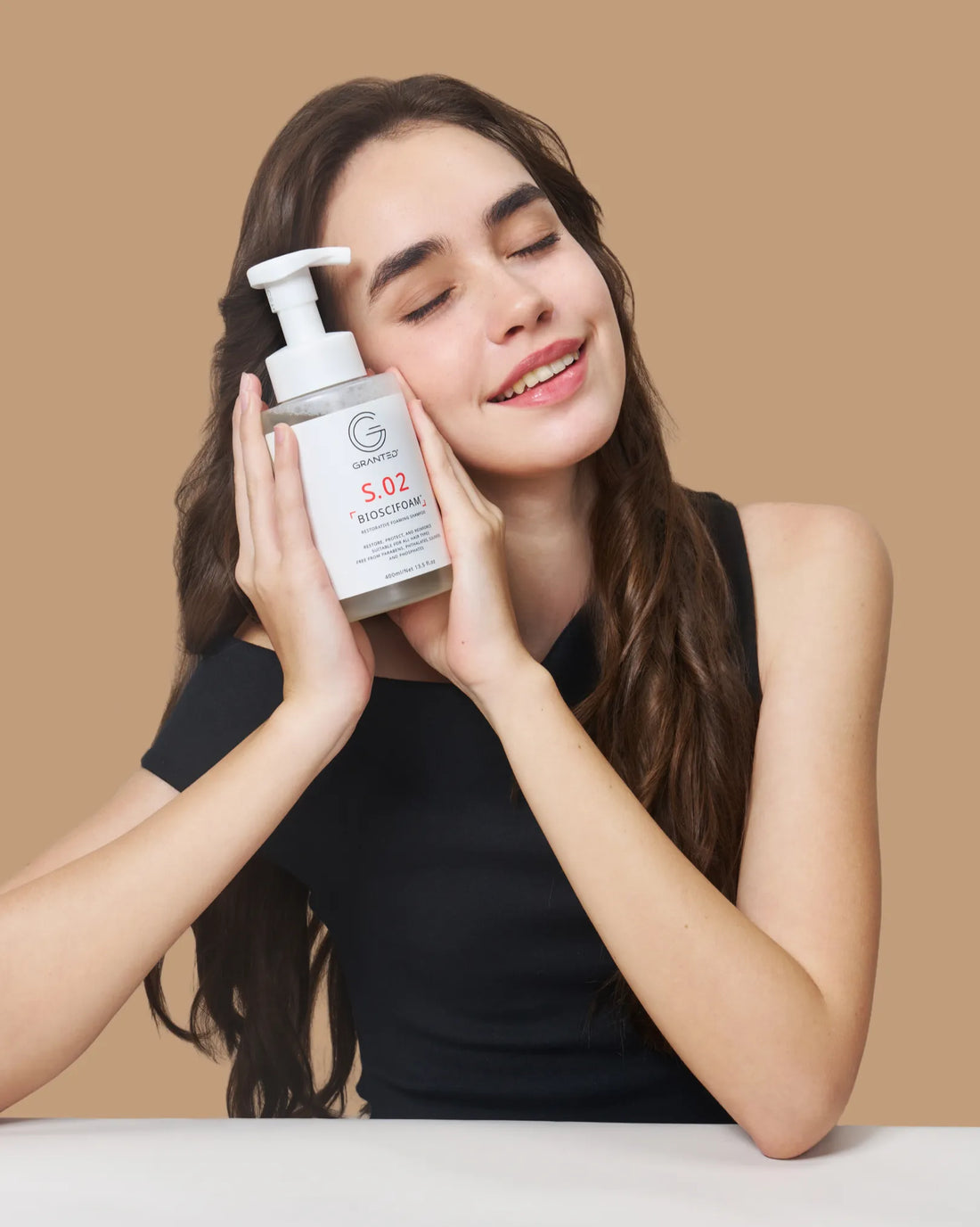 Model holding Granted S.02 BIOSCIFOAM repair shampoo to her cheek, for damaged hair.